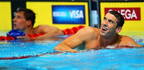 Michael Phelps sorri após vitória, enquanto Lochte, ao fundo, observa o placar