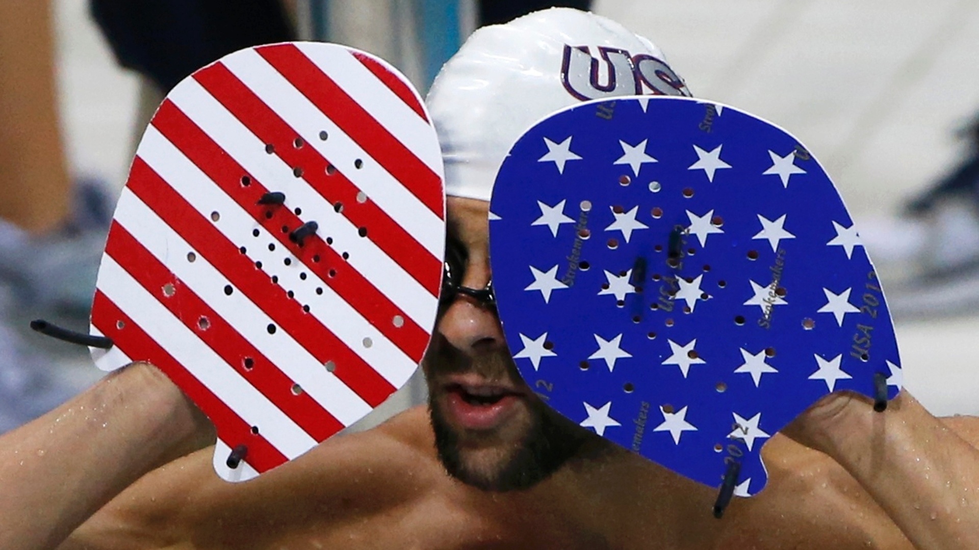 Michael Phelps tenta remover seus óculos de natação enquanto usa equipamento nas cores da bandeira dos EUA (24/07/2012)