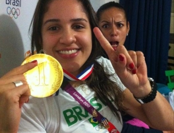natália, da equipe brasileira de vôlei feminino, faz uma montagem como se estivesse usando a medalha de ouro em troca da credencial; ao fundo, vemos Paula Pequeno.