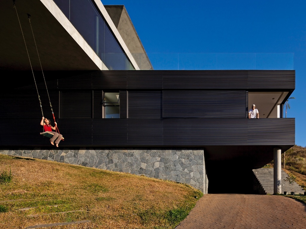 Um balanço de brinquedo foi instalado sob a laje do bloco social, em balanço construtivo, da casa EG. O projeto da residência em Nova Lima (MG) é assinado pelo arquiteto Marcelo Alvarenga. Na fachada, destaque para as pedras usadas no embasamento, para as venezianas de alumínio anodizado, além da massa de cimento e dos panos de vidro - Leonardo Finotti/ UOL