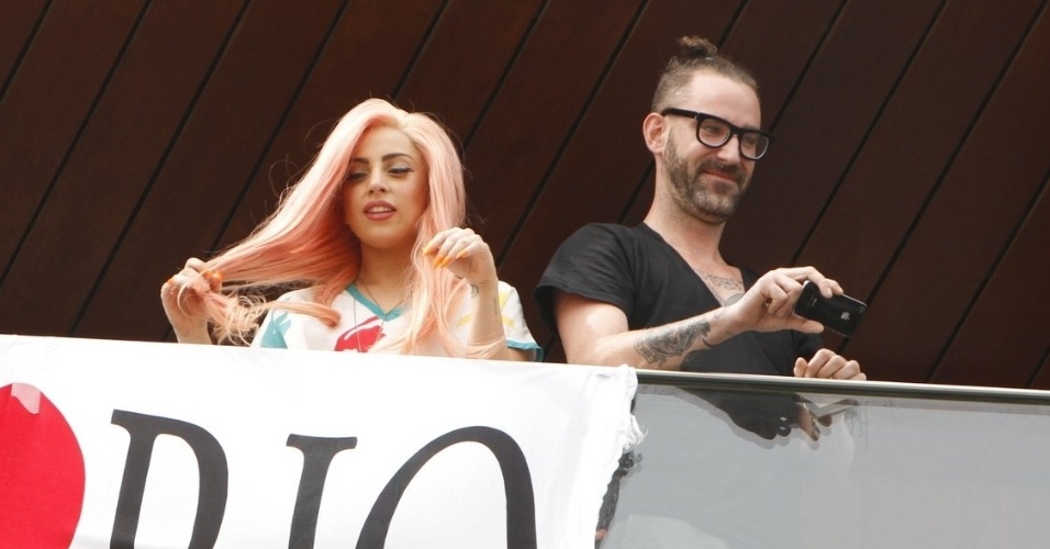 Após sair na sacada do hotel com a camisa da seleção brasileira de futebol, Lady Gaga retorna usando um vestido branco. Ao lado da bandeira "I Love Rio", ela acena e manda beijos para os fãs cariocas (8/11/12)