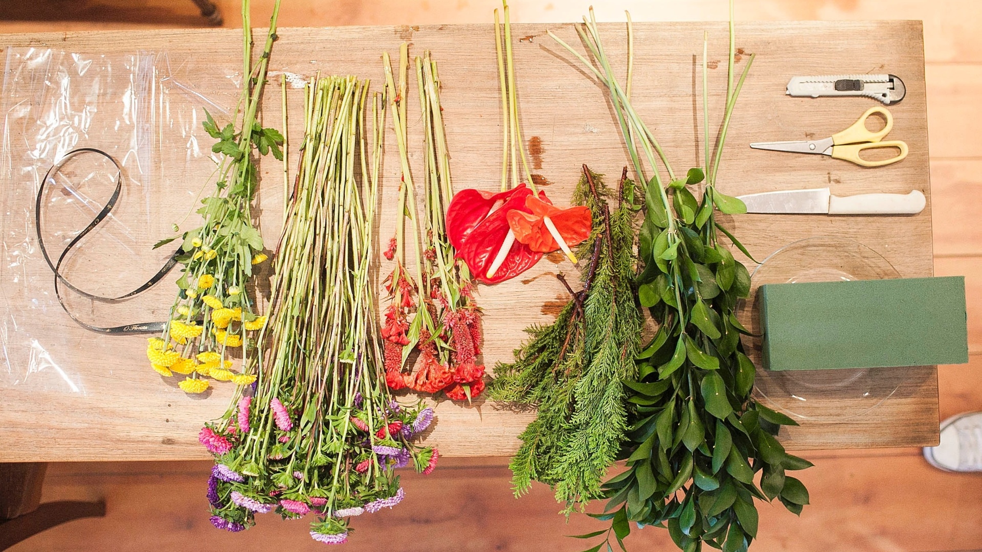Materiais: papel celofane / fita de cetim ou tecido / flor pinóquio (dura duas semanas) / flor rainha margarida (dura duas semanas) / flor crista de galo (dura uma semana) / flor antúrio (dura uma semana e meia) / tuia (dura uma semana) / ruscus (dura três semanas) / estilete / tesoura / faca / espuma floral / água / bancada que possa ser molhada / prato ou tábua - Leonardo Soares de Souza/ UOL