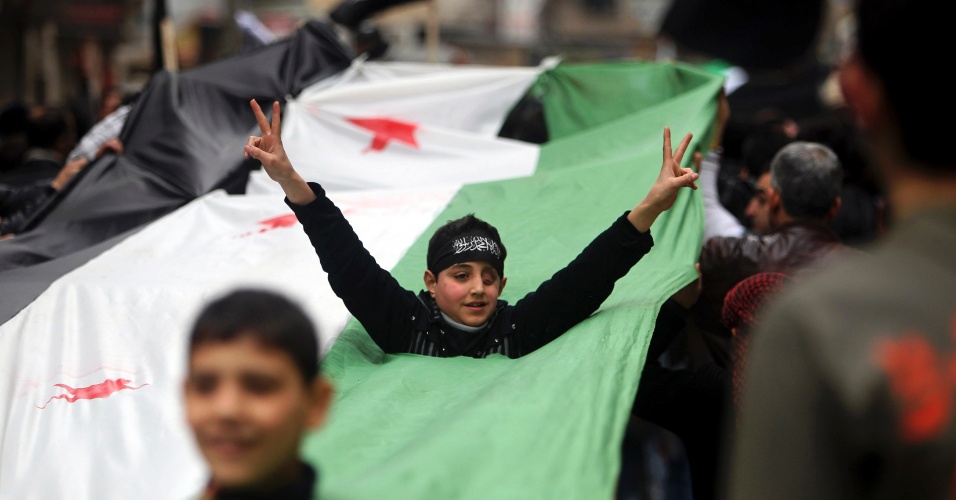 22.fev.2013 - Garoto levanta os braços através de um buraco numa bandeira da oposição síria, durante protesto contra o presidente Bashar Assad, em Aleppo