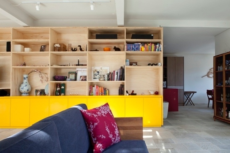 Os arquitetos Tito e Chantal Ficarelli, do escritório Arkitito, optaram por aplicar a tonalidade amarela vibrante somente na parte inferior da estante. Outras cores compõem o ambiente de estar: o azul do sofá e o tom rosa da almofada - Divulgação