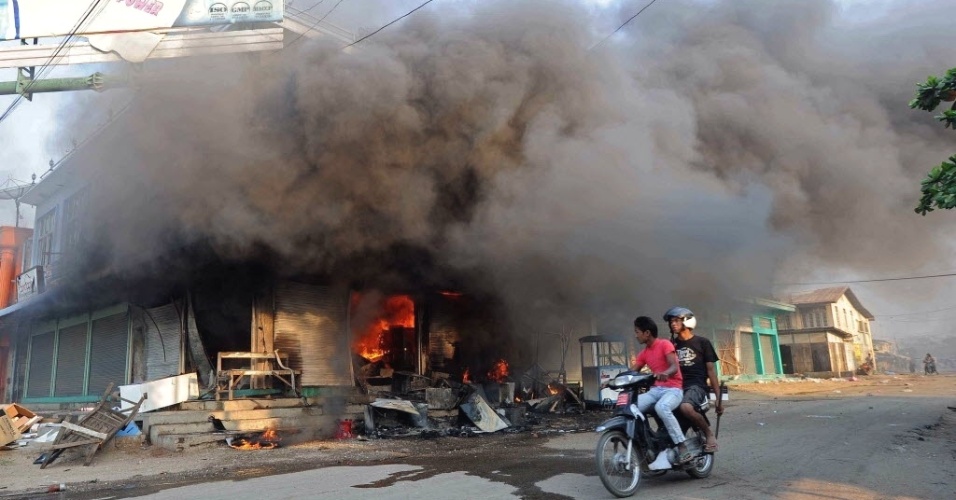 22.mar.2013 - Moradores circulam de moto em frente a edifício em chamas em área de Miktila, região central de Mianmar, onde os confrontos religiosos entre budistas e muçulmanos já deixaram pelo menos 20 mortos
