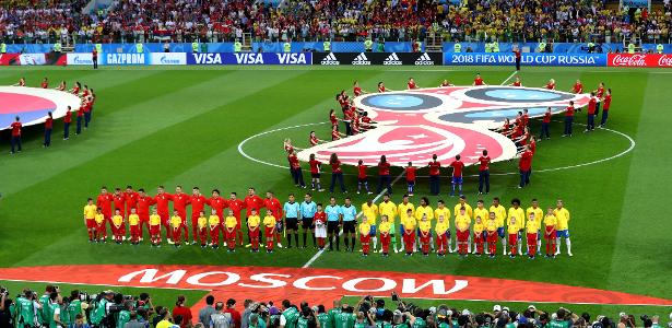 A wide shot of a football pitch with players in national team kits in the center circle during a pre-match ceremony, under dramatic stadium lighting.