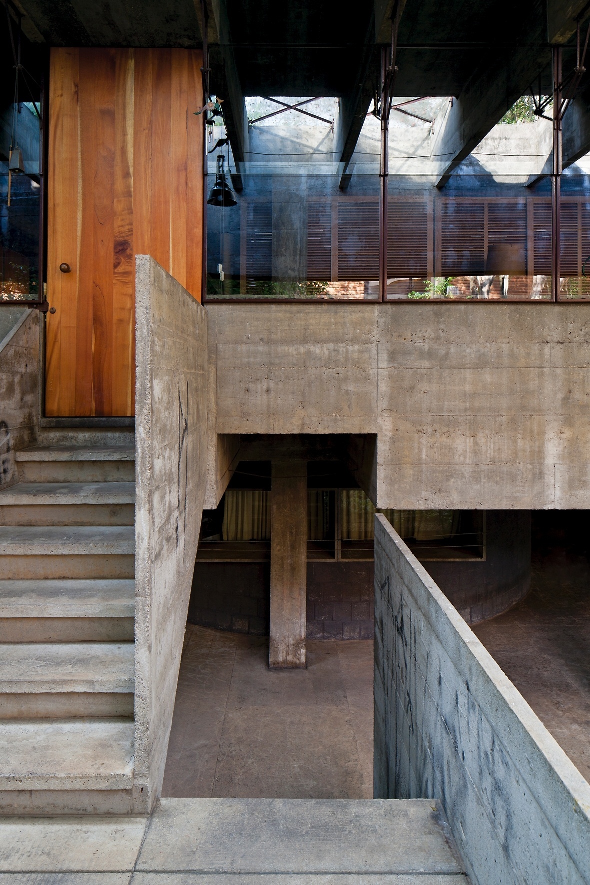 Na escada de acesso ao pavimento superior está um dos poucos elementos de madeira da casa: a porta de entrada. No piso inferior, destaque para um dos quarto pilares que suportam toda a construção. A superfície envidraçada reflete o beiral constituído por um pergolado, projetado pelo arquiteto Paulo Mendes da Rocha - Leonardo Finotti/ UOL