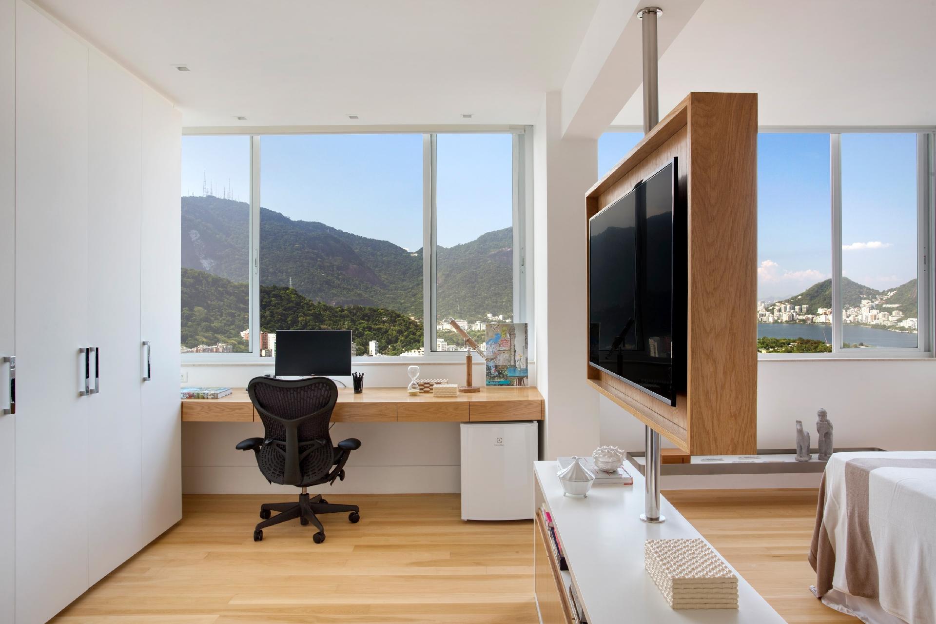 Este apartamento conta com uma vista aproveitada pela RBP Arquitetura e Interiores ao posicionar o home office integrado ao quarto de casal. Eles desenvolveram uma marcenaria com painel pivotante que possibilita assistir a TV de todos os ângulos dos ambientes. Para quem não abre mão de ter tudo organizado, lançar de mão de gavetas e uma bancada espaçosa é essencial - Juliano Colodeti / MCA Estudio | Divulgação