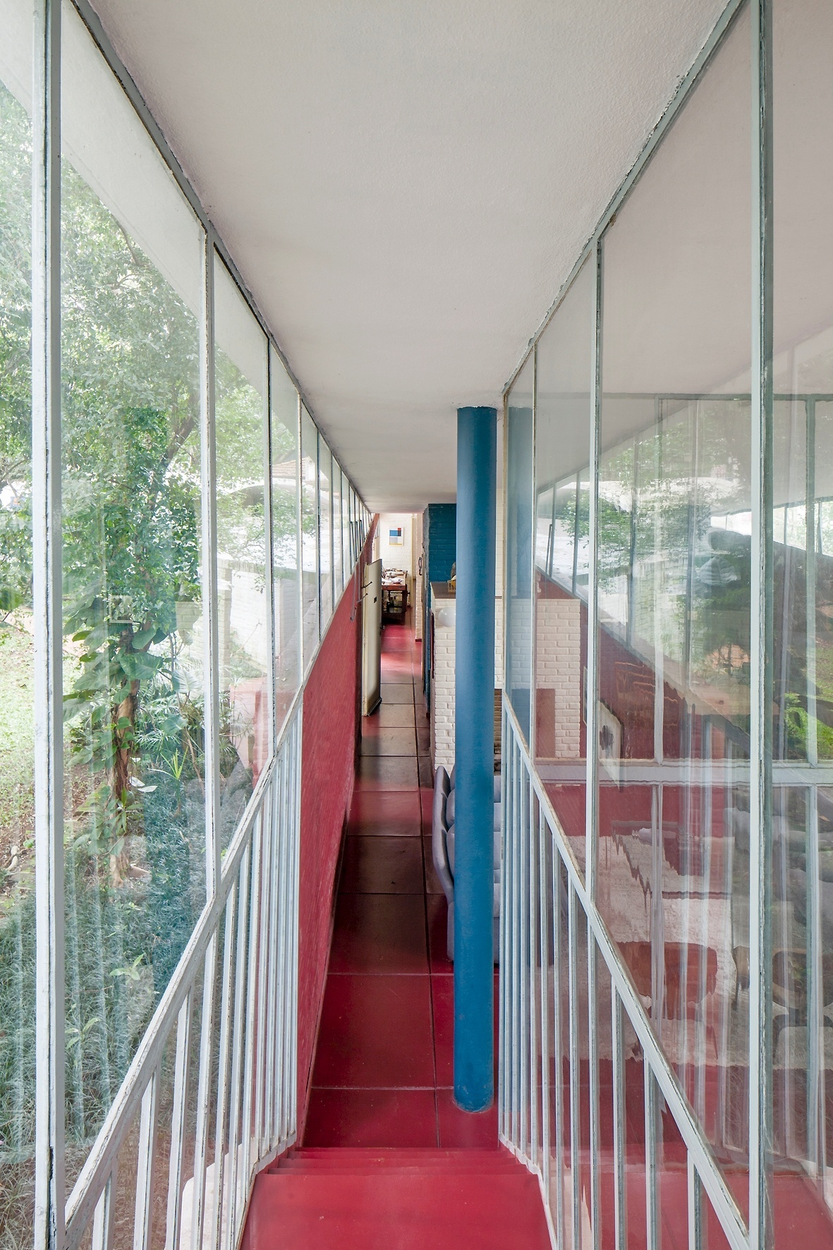 Do alto da escada que liga o térreo ao mezanino, onde está o estúdio, é possível visualizar o extenso corredor que percorre os ambientes sociais (estar com lareira e jantar), além de cozinha, banheiros e, ao fundo, um dos dormitórios. Os fechamentos em vidro garantem a transparência necessária para entrada de luz natural e integração com o exterior. A Casa do Arquiteto foi projetada por João Batista Vilanova Artigas, em 1949 - Leonardo Finotti/ UOL