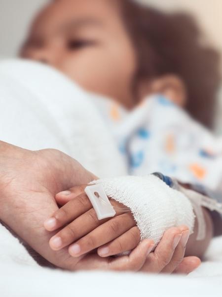 Criança em cama de hospital segurando a mão de um adulto - Sasiistock/Istock