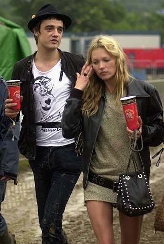 Nos bastidores do festival Glastonbury em 2005, o músico inglês Pete Doherty caminha com a então namorada Kate Moss. Ele usa chapéu fedora no melhor estilo roqueiro com jaqueta de couro, camiseta e jeans - Getty Images