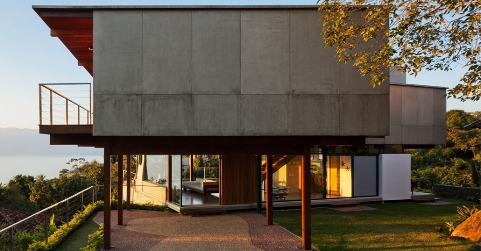 Deste ângulo é possível observar que o volume do pavimento superior está parcialmente apoiado em pilotis na casa Embaúba. As superfícies "cegas" são constituídas por painéis de placa cimentícia. O projeto da casa de praia, assinado por Flavia Cancian, venceu na categoria "Residencial - Praia" o prêmio "O Melhor da Arquitetura", promovido pela revista Arquitetura & Construção, em 2012 - Nelson Kon/ UOL