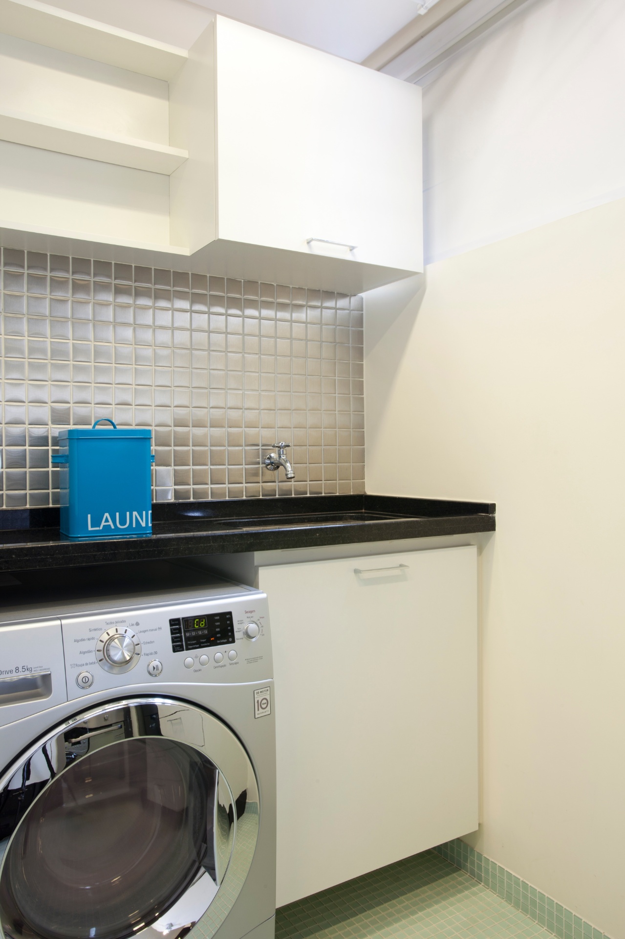 Área de serviços pôde ser bem aproveitada, depois da diminuição do banheiro (ao lado), pois nela não havia originalmente espaço para a máquina de lavar roupas. Os revestimentos acompanham os da cozinha: pastilhas aço inox (Mozaik), nas paredes, e de vidro (Colormix), no piso. A bancada é de granito preto (Duarte Mármores e Granitos) e os móveis foram executados pela Madero Marcenaria em MDF laqueado. O projeto de reforma é da designer de interiores Adriana Fontana - Divulgação