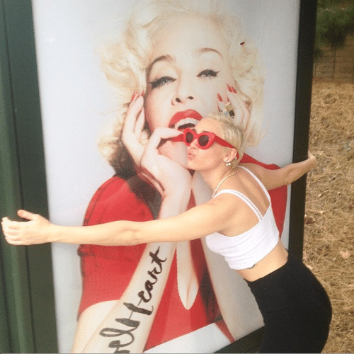 12.mar.2015 - Miley Cyrus abraça e tira foto com imagem de Madonna na propaganda do novo álbum da diva pop