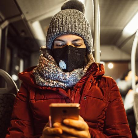 mulher lendo celular no ônibus, máscara, coronavírus, fake news - iStock