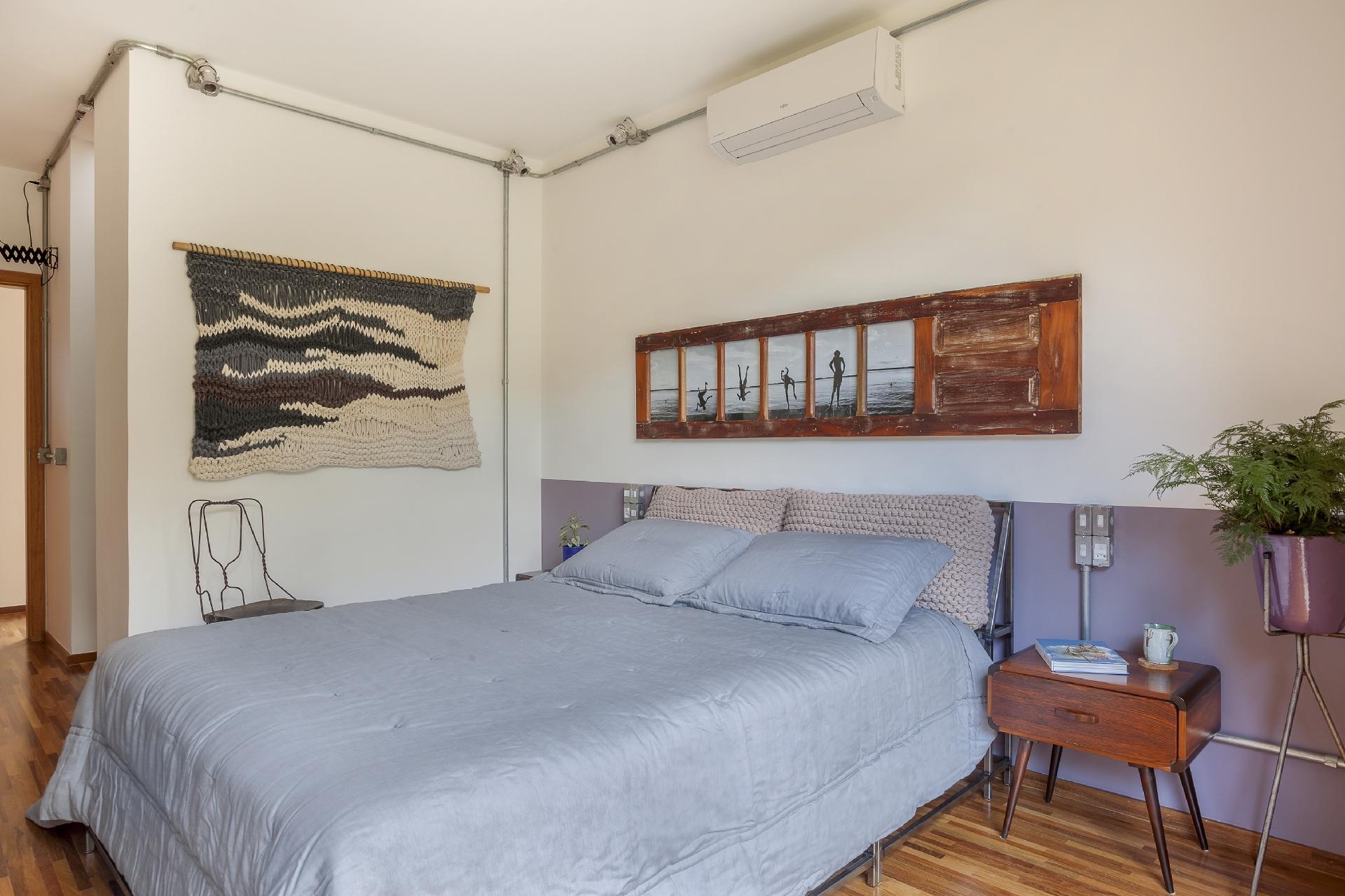 A pintura da meia parede na cor lilás e na altura da cabeceira serviu para destacar a estilosa estrutura de metal em aço carbono da cama (Prototype). Marca dos projetos do arquiteto Fabio Marins, o reaproveitamento de materiais se dá neste quarto por meio da restauração da porta de demolição sobre a cama (Degar), que se transformou em um lindo painel de fotos para a moradora fotógrafa - Alessandro Guimarães/ Divulgação