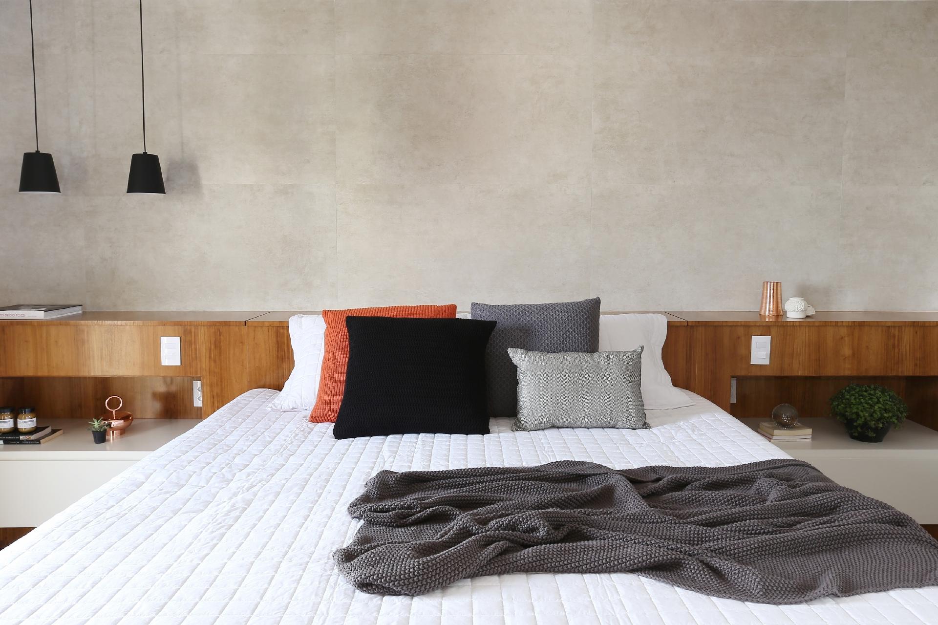 A marcenaria que forma a cabeceira esconde um baú, que guarda os edredons. Feita de MDF com folha de madeira cabreúva parda, ela traz ainda dois criados-mudos embutidos na cor cinza, que contrastam com o tom quente da madeira e combinam com a parede de porcelanato cimentado logo atrás. A composição traz aconchego com um toque de urbanidade. Projeto Degradê Arquitetura e Interiores - Mariana Orsi/ Divulgação