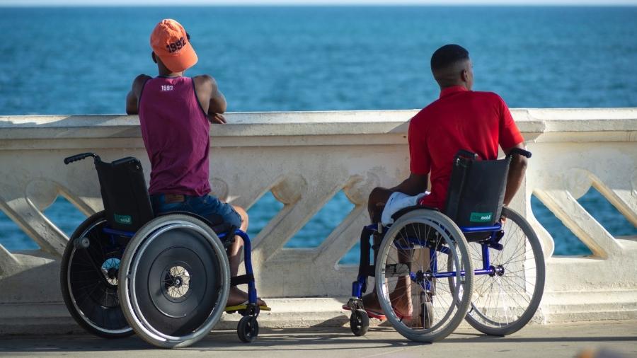 Dois jovens em cadeiras de rodas olhando para o mar. - iStock
