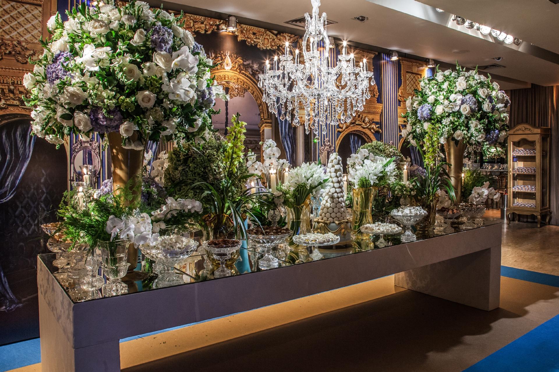 Os espelhos e as peças de cristais embelezaram a mesa do bolo da debutante. Orquídeas, rosas, lisianthus e boca de leão compunham os arranjos dispostos na mesa e confeccionados pelo florista Andre Pedrotti - Lan Rodrigues