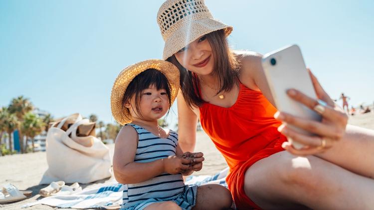 Mãe tirando foto com a filha na praia/ Mãe brincando com filha na areia/ Mãe fotografando a filha na areia