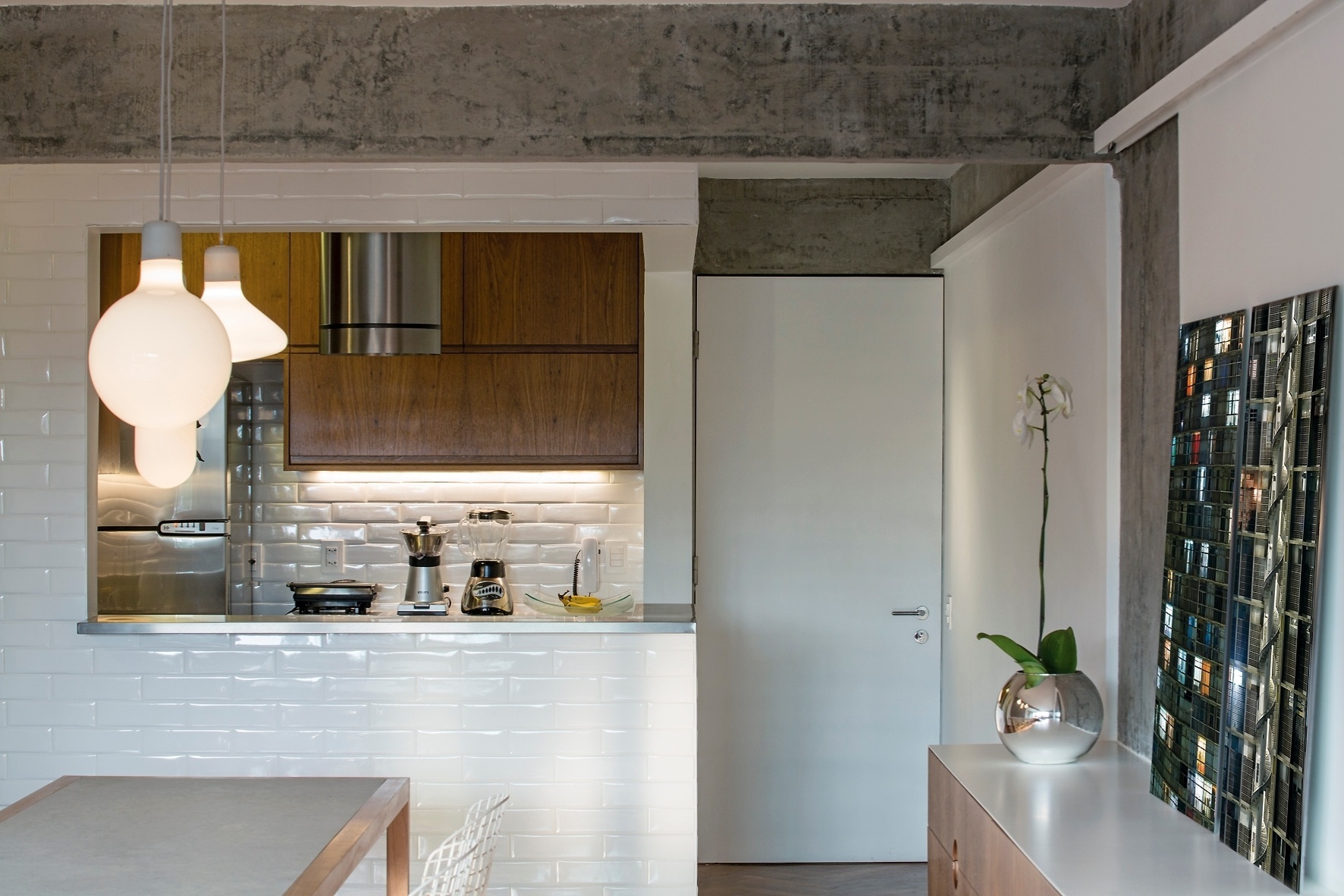 A cozinha do Apartamento FT tem passa-pratos voltado para o jantar, com bancada em aço-inox (Mekal) e paredes revestidas com azulejos cerâmicos brancos. Calhas metálicas em aço-carbono (DIX Arte&Metal) contornam a área social e embutem a iluminação indireta, próxima ao forro. Sobre a mesa, destaque para os pendentes leitosos. A reforma do apê dos anos 50 foi projetada pelo escritório Pascali Semerdjian Arquitetos - Leonardo Finotti/ UOL