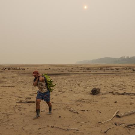 Agricultor caminha carregando cacho de banana por faixa de areia que se formou no rio Madeira, em Porto Velho (RO)