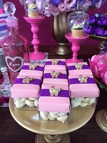 Essas caixinhas fofas servidas no chá de fraldas eram de acetato e estavam recheadas com sequilhos. O acabamento usa fita de gorgurão roxa e aplique de carrinho de bebê dourado.  Ao fundo, dois porta-cupcakes da Alugue para Festas exibem minicupcakes de chocolate com ganache - Divulgação
