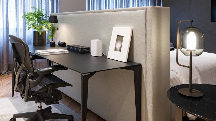 No quarto do casal, o escritório é integrado ao quarto de forma criativa, ideia da arquiteta Consuelo Jorge (https://www.instagram.com/consuelojorge/). Apoiado na cabeceira da cama, de tecido, o office escuro segue a mesma linguagem de todo o amplo cômodo, onde a pintura especial de concreto no forro garante o ar despojado e contrasta com piso de madeira, que aquece e dá conforto. Atrás da mesa, há uma grande estante com objetos e peças de design garimpados pelos moradores. - Divulgação - Divulgação