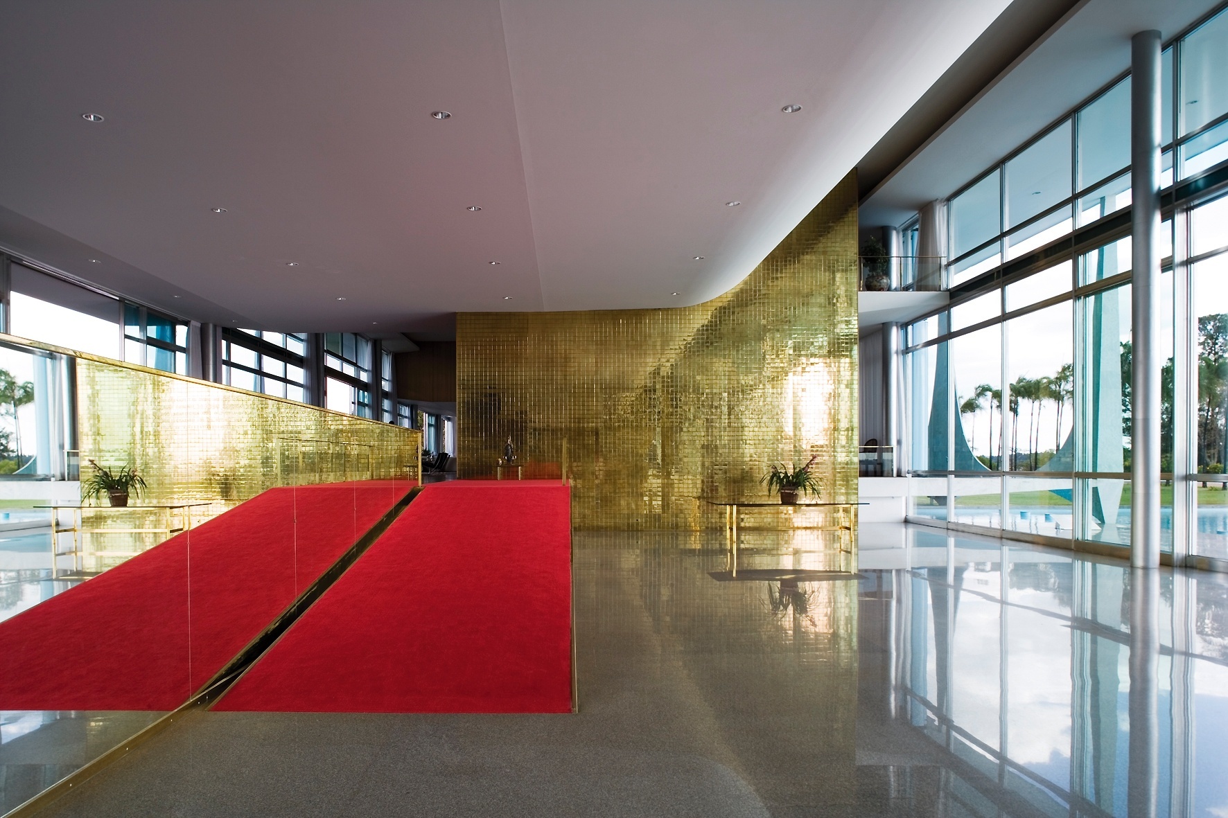 A monumentalidade do hall de entrada do Palácio da Alvorada é marcada pelo pé-direito duplo e pelo grande painel com placas de latão dourado (ao fundo), assinado por Athos Bulcão. Destaque também para a rampa revestida pelo tapete vermelho, ao lado da parede espelhada. O projeto de arquitetura é de Oscar Niemeyer - Leonardo Finotti/ UOL