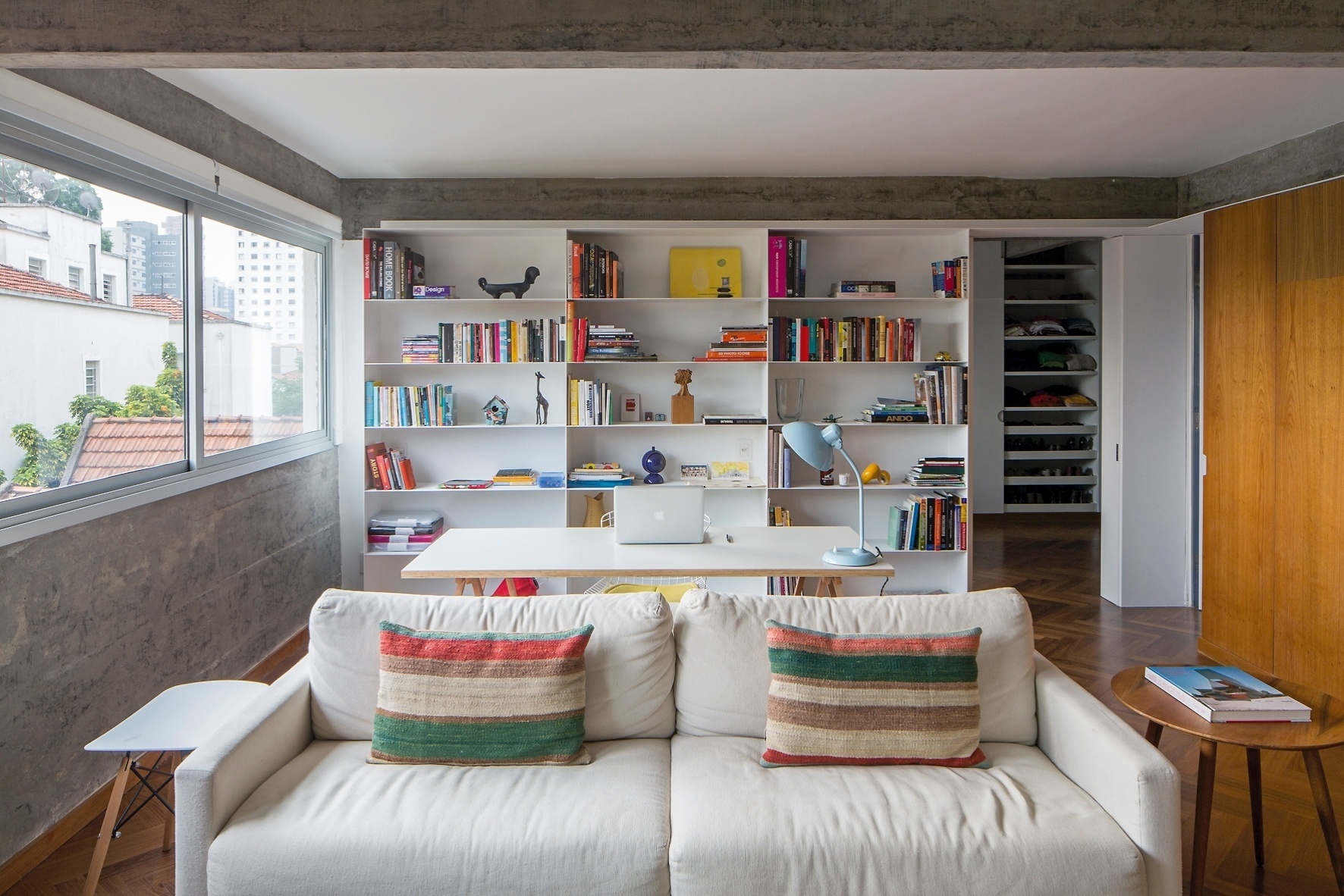 O espaço de trabalho entre o estar com TV e o dormitório (ao fundo) conta com nichos feitos com chapas de aço-carbono pintadas de branco (DIX Arte&Metal). À direita, uma chapa avança sobre a porta do dormitório, contornando a parede para emoldurar um armário. A ideia dos projetistas do escritório Pascali Semerdjian era encaixar a marcenaria nesses módulos metálicos - Leonardo Finotti/ UOL