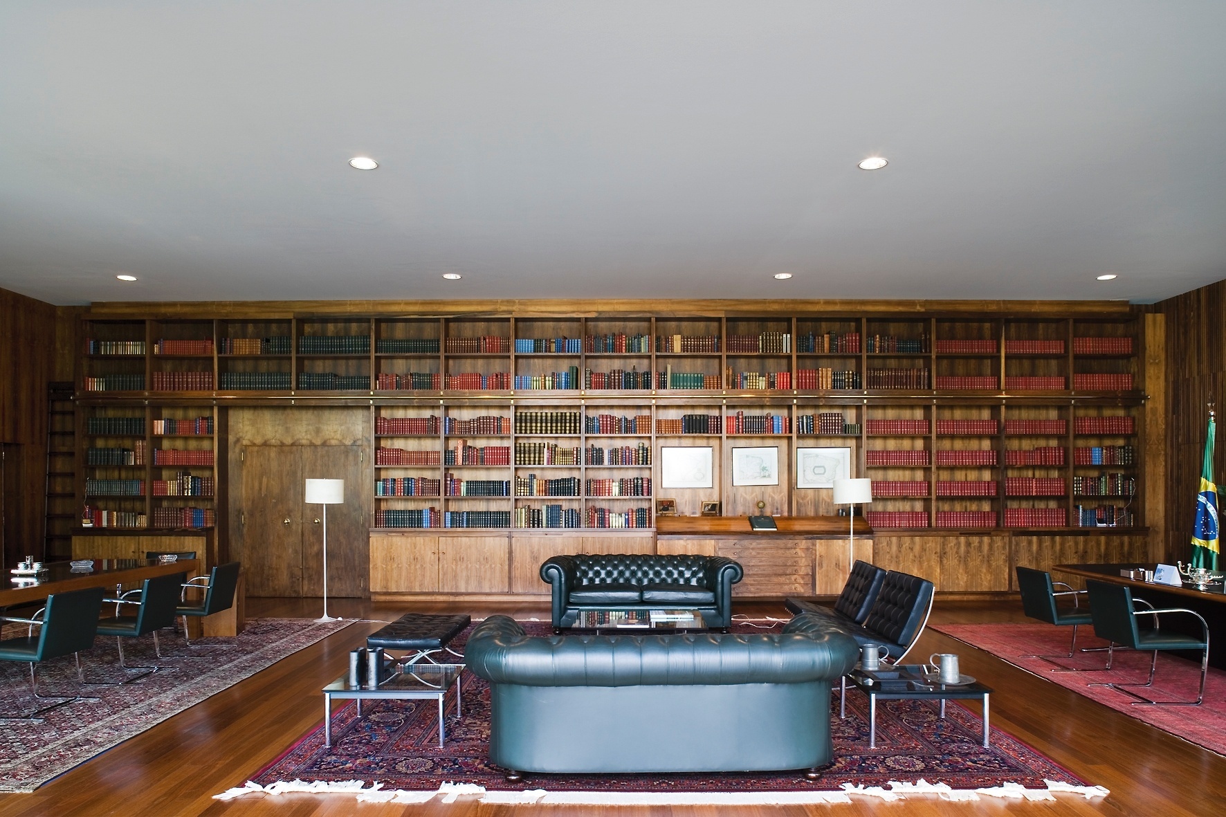 Um dos ambientes mais suntuosos do Palácio da Alvorada, concebido pelo arquiteto Oscar Niemeyer, a biblioteca conta com estantes e paredes revestidas com jacarandá. O mobiliário é composto de peças clássicas como  os sofás Chesterfield e exemplares modernos como as cadeiras e bancos Barcelona e as cadeiras Brno (junto às mesas, na laterais), criados por Mies Van der Rohe. No chão, generosos tapetes persas e, nas estantes, obras de escritores como Manuel Bandeira e Carlos Drummond de Andrade - Leonardo Finotti/ UOL