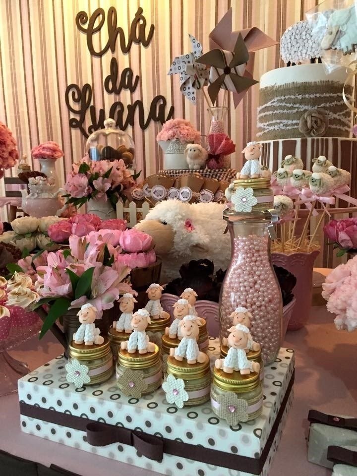 Caixa de MDF forrada com tecido. Sobre ela, potinhos de doce de amendoim decorados com ovelhinha, leiteira de vidro com pérolas rosa e regador em miniatura servindo de vaso para as astromélias naturais. Ao fundo, vaso de louça com cakepops (bolo no palito) de chocolate, decorados como ovelhinhas, e cones de papel kraft com bombons de biscoito wafer. Tudo para deixar o chá de fraldas mais elegante - Divulgação