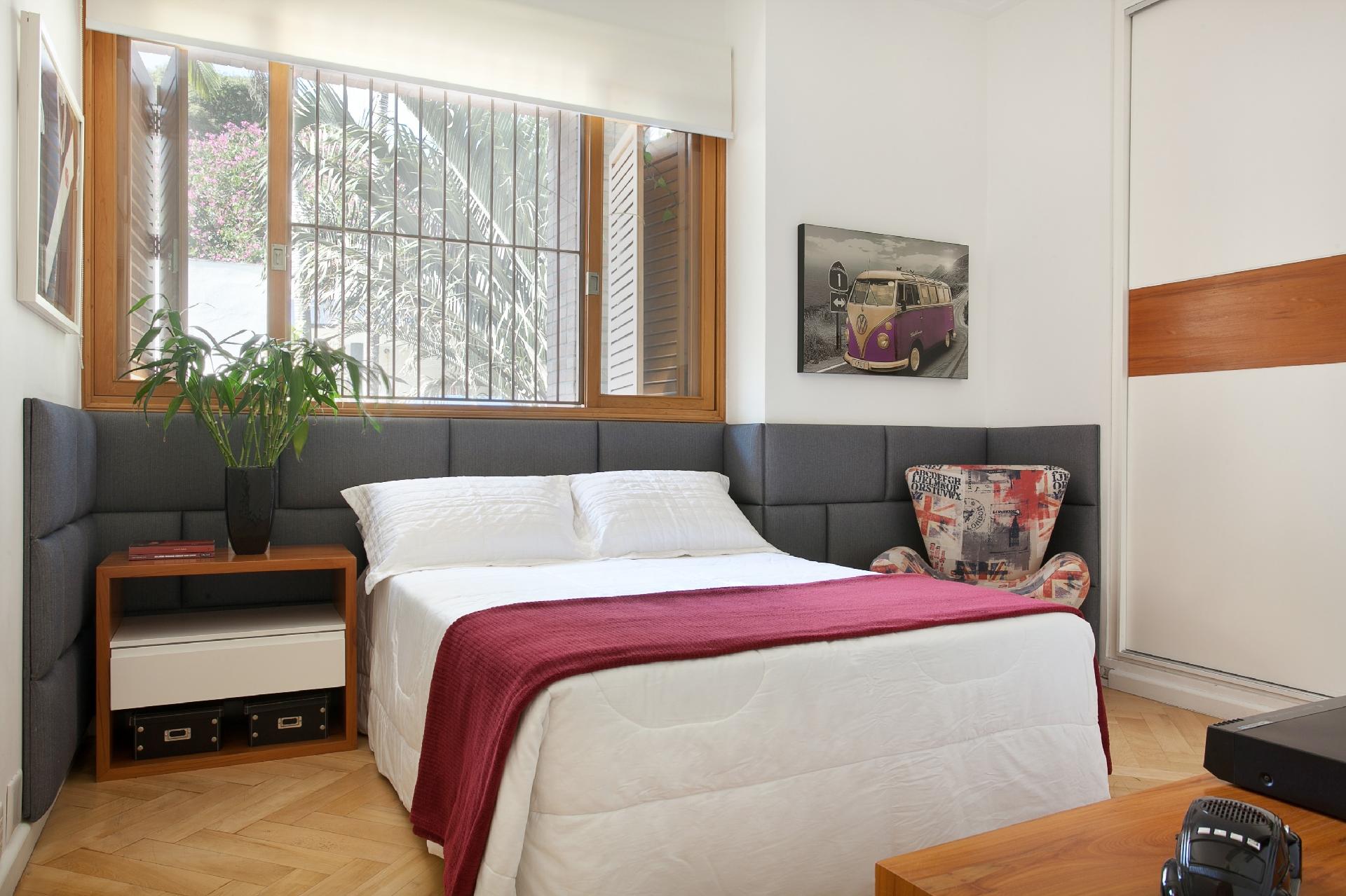 A cabeceira de seda mista foi construída em placas almofadadas que circundam parte da parede como se abraçasse a cama. A extensão dela, segundo a arquiteta Beatriz Quinelato, deixa o quarto ainda mais amplo e cria uma sensação acolhedora. A neutralidade do cinza e da parede branca permite uma roupa de cama mais colorida e não briga com a estampada poltrona ao lado - Adriana Barbosa/ Divulgação