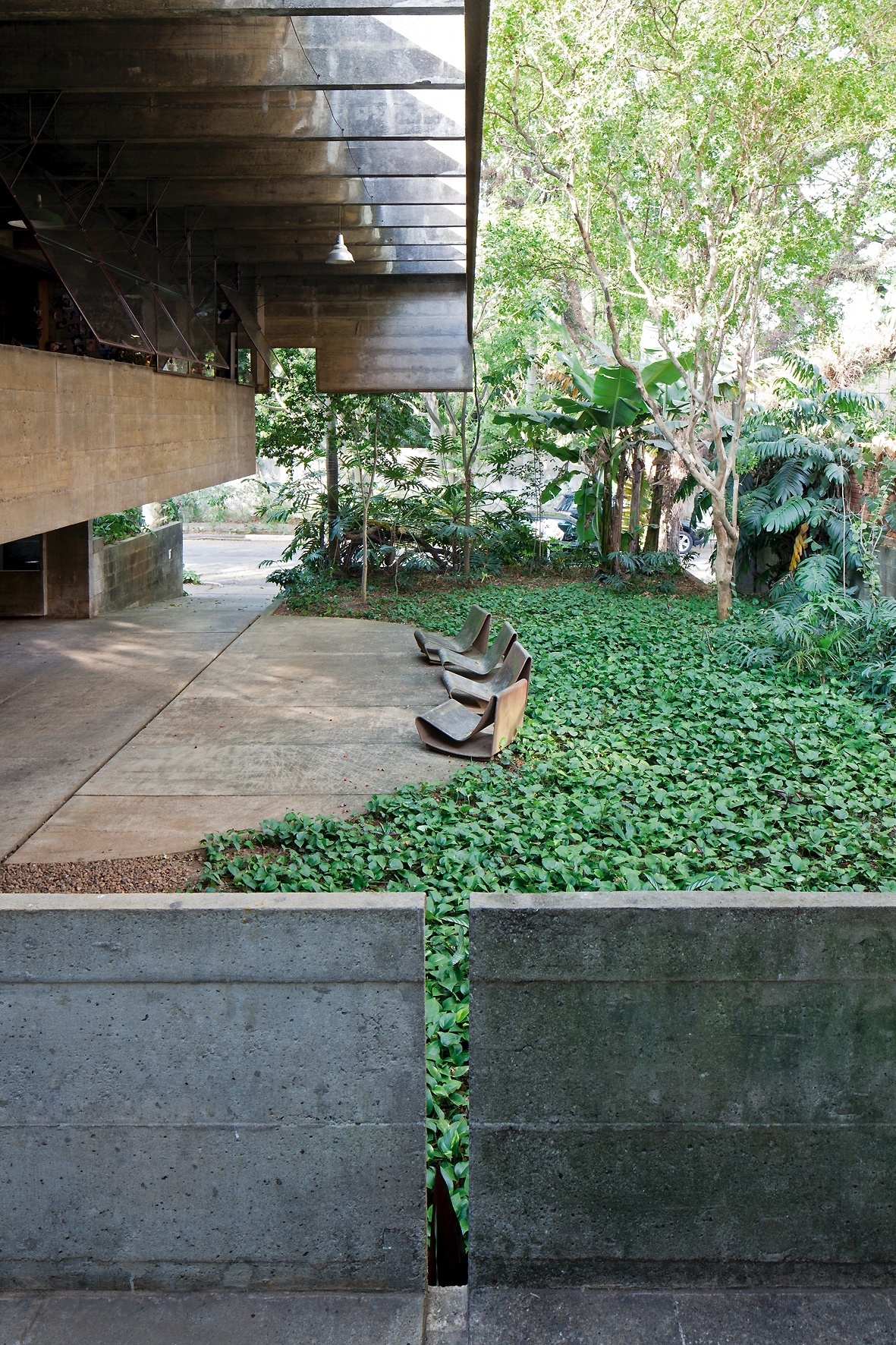 Em primeiro plano, o guarda corpo em concreto apresenta uma abertura usada para o escoamento da água da chuva. O jardim com vegetação intensa quebra a hegemonia do concreto e forma uma área agradável com vista para a rua. A casa Butantã foi projetada pelo arquiteto Paulo Mendes da Rocha nos anos 1960 - Leonardo Finotti/ UOL