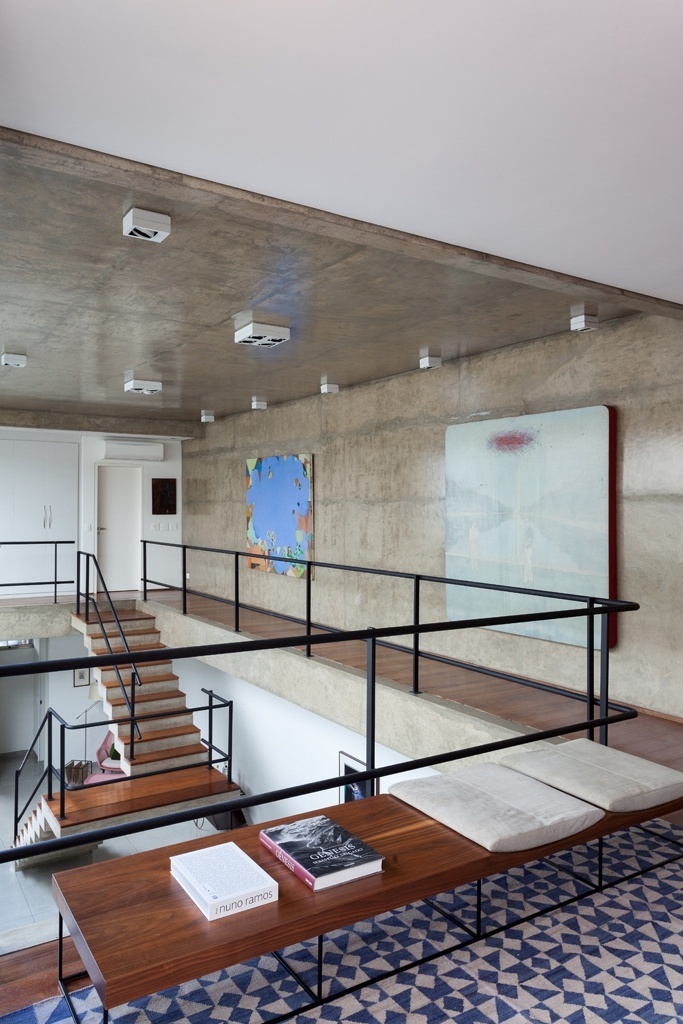 A partir da sala íntima do pavimento superior observa-se o pé-direito duplo e as paredes e teto em concreto aparente contrastando com a madeira do piso, dos degraus da escada e do banco BA-102, da Etel. O tapete azul e branco é da By Kamy. A Casa Jardins foi projetada pelo escritório CR2 Arquitetura - Fran Parente/ Divulgação