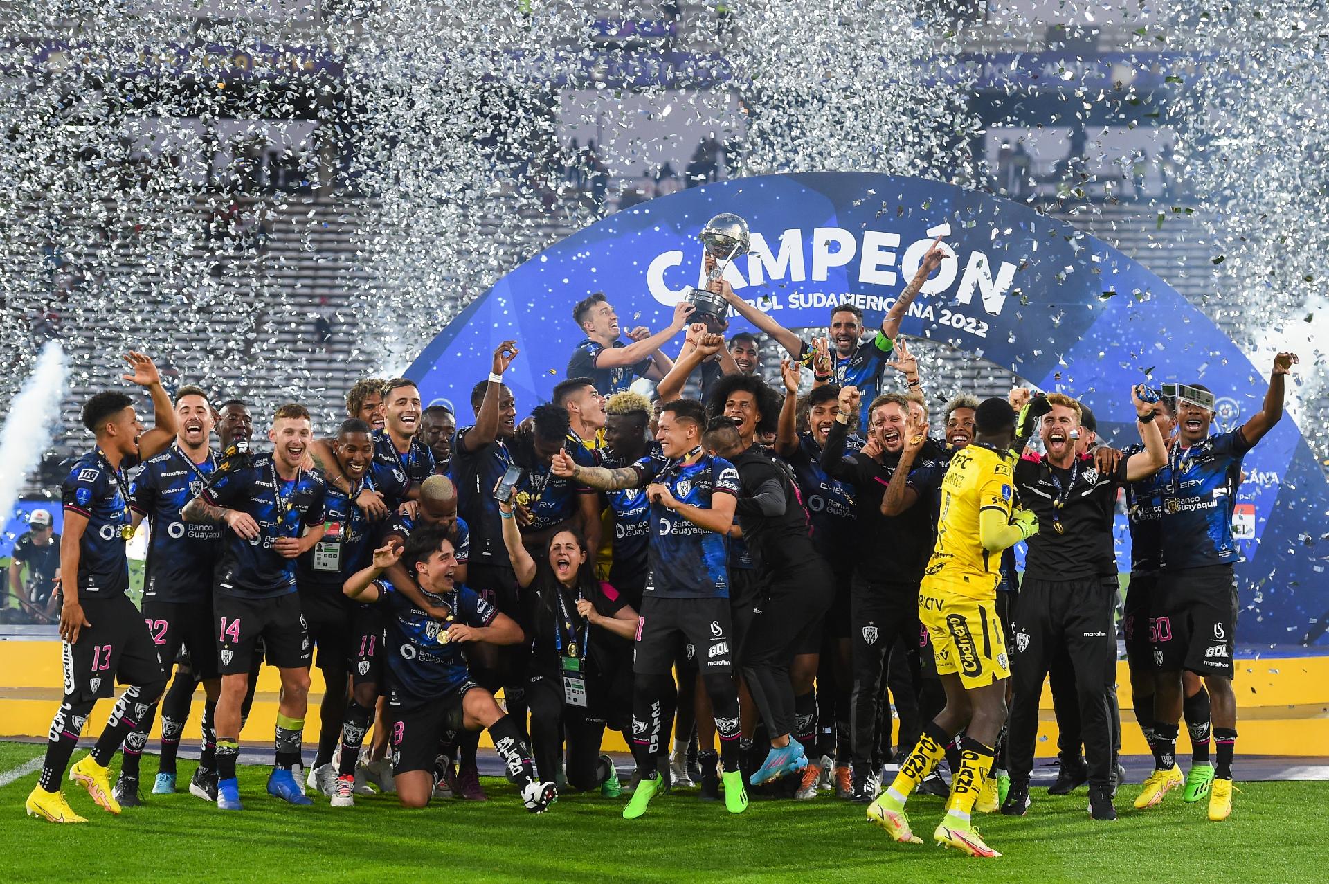 Jogadores do Del Valle festejam título da Copa Sul-Americana de 2022. Imagem mostra festa, com confetes prateados no ar e, ao fundo, taça sendo levantada por três jogadores. 