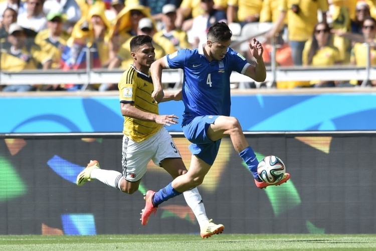 Jogador grego, Konstantinos Manolas, parece voar para escapar de marcação de colombiano durante partida de estreia das equipes na Copa