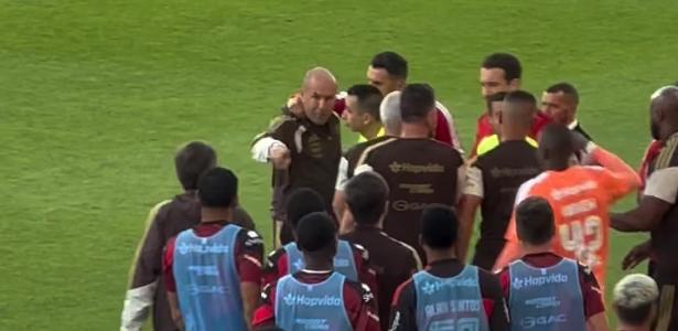 Zwei Fußballtrainer im Gespräch, angespannte Atmosphäre durch dramatische Beleuchtung und gegenüberliegende Spielfeldseiten angedeutet.