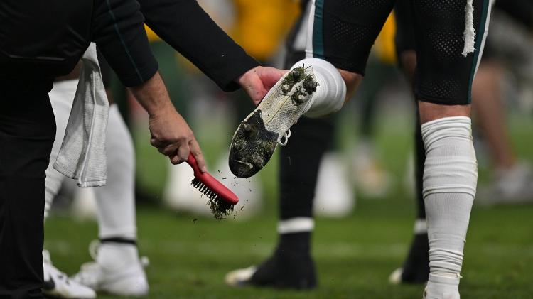 Funcionário do Philadelphia Eagles tira grama da chuteira de jogador durante jogo da NFL no Brasil