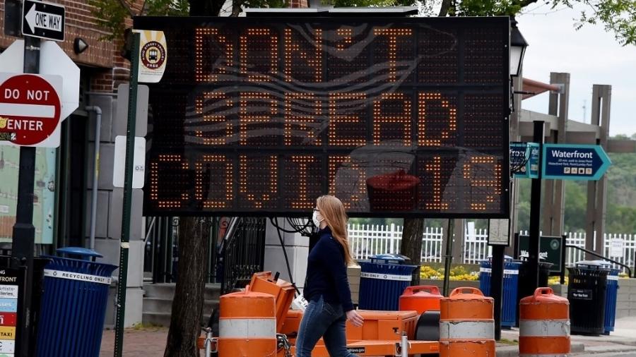 Mulher usando máscara caminha sob placa com os dizeres: "Não espalhe covid-19" em rua de Alexandria (Virgínia, EUA) - Olivier Douliery/AFP