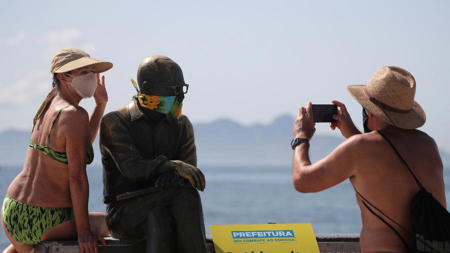 Uma mulher posa para uma foto ao lado da estátua do escritor brasileiro Carlos Drummond de Andrade, que usa uma máscara protetora no primeiro dia de uso obrigatório de máscaras na cidade do Rio de Janeiro, em abril de 2020 - SERGIO MORAES/REUTERS