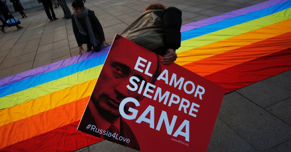 5.fev.2014 - Um manifestante ajuda a exibir uma bandeira de arco-íris, símbolo do movimento LGBT, quanto segura um cartaz mostrando o presidente russo Vladimir Putin e a frase 