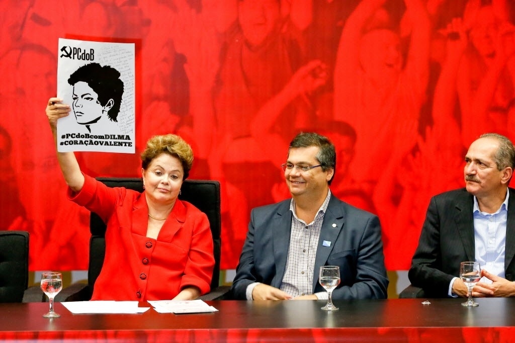 27.jun.2014 - A presidente Dilma Rousseff ergue cartaz com a foto dela feita na ocasião de seu julgamento pelo tribunal militar na época da ditadura, onde escreveu uma mensagem destinada à UJS (União da Juventude Socialista), durante a convenção nacional do PC do B, nesta sexta-feira (27). Na ocasião, o partido ratificou o apoio à reeleição da presidente. Ao lado de Dilma estão o candidato ao governo do Maranhão Flávio Dino (esq.), e o ministro do Esporte, Aldo Rebelo (dir) - Pedro Ladeira/Folhapress