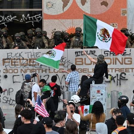 10.jun.2025 - Manifestantes agitam bandeiras do México enquanto policiais protegem um prédio federal em Los Angeles 10.jun.2025 - Manifestantes agitam bandeiras do México enquanto policiais protegem um prédio federal em Los Angeles