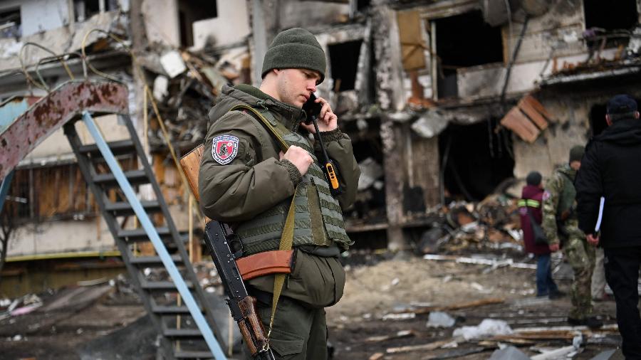 Soldado ucraniano fala em um smartphone em frente a um prédio residencial danificado em um subúrbio da capital ucraniana Kiev, atingido por um projétil militar russo - Daniel Leal/AFP