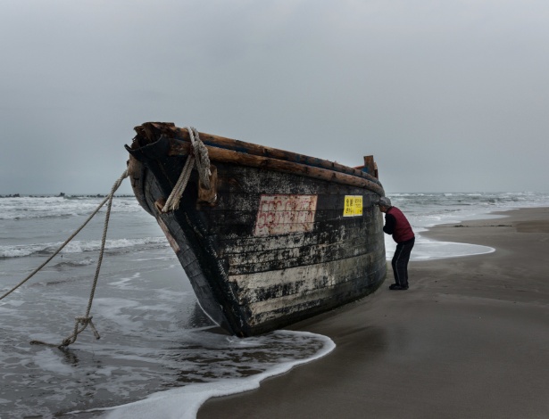 Homem tenta espiar lado de dentro de um barco de madeira que desembarcou com oito cadáveres na praia de Miyazawa, no noroeste do Japão - KO SASAKI/NYT