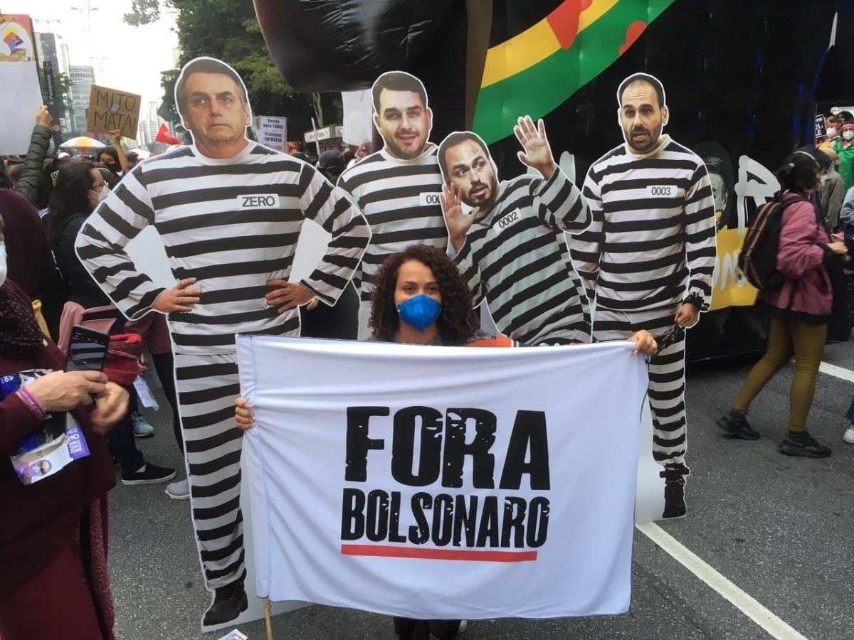 Manifestante posa com faixa em frente a cartazes que simulam Jair Bolsonaro e seus filhos com roupas de presidiário - Amanda Rossi/UOL