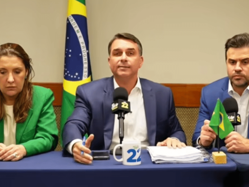 Flávio Bolsonaro ao centro de uma mesa, com microfone e aliados, durante transmissão ao vivo.
