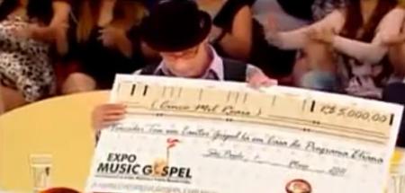 Cheque figurativo que foi dado para vencedora do concurso; na imagem jurado Arnaldo Saccomani segura o prêmio