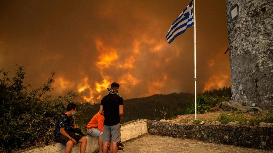 Moradores da ilha de Eubeia observam o avanço das chamas em um mirante com a bandeira da Grécia - Angelos Tzortzinis/AFP